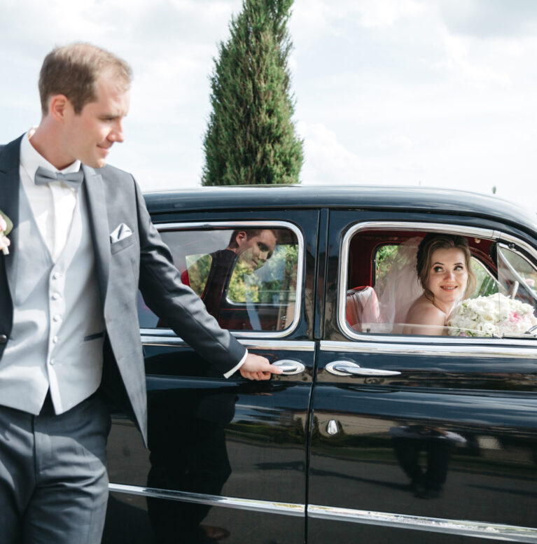 The bridegroom is standing by the car while the bride is in the car