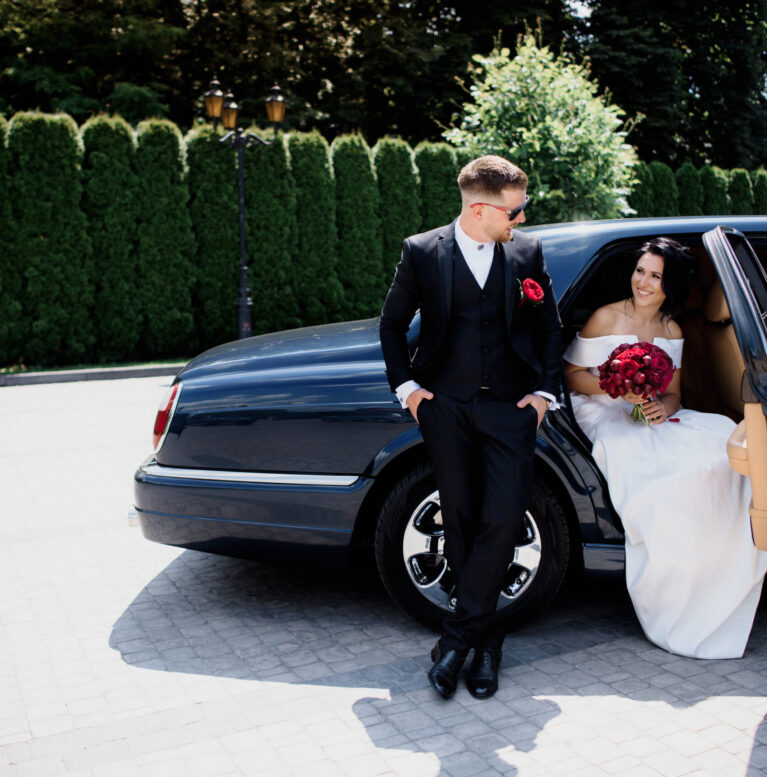 Beautiful wedding couple is smiling in the black car on the sunny day, dressed in elegant wedding outfits with red bouquet