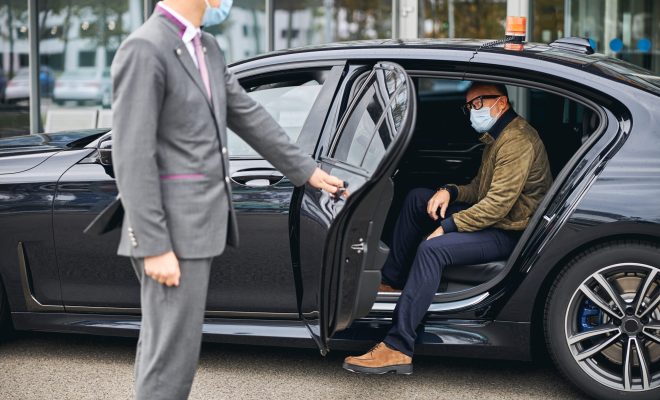 Chauffeur opening the car door for a Caucasian male passenger in a disposable face mask